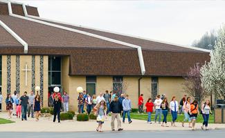 A group of people, including men, women, and children, exit a modern church building with brown roof and cross symbol in an outdoor setting. Trees and shrubs decorate the surroundings.